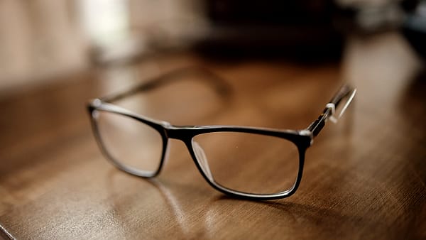A pair of black eyeglasses resting on a wooden desk, with daylight coming through a window nearby