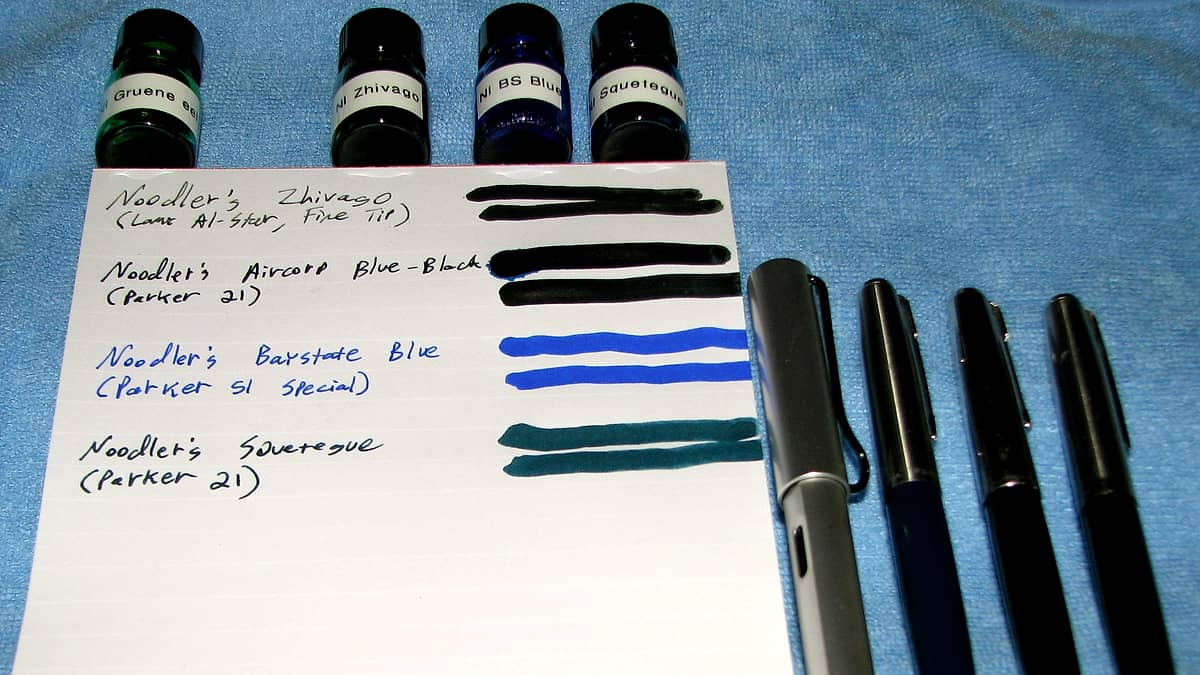 A blank A5 writing pad on a desk beside a row of ink pens laid out in a sampler tray