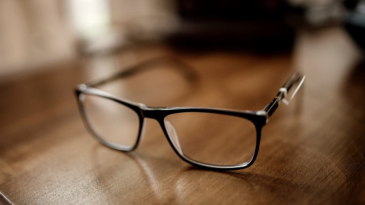 A pair of black eyeglasses resting on a wooden desk, with daylight coming through a window nearby
